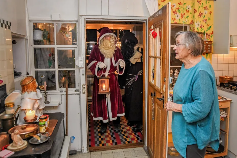 Samichlaus und Schmutzli stehen mit Laterne in der Küchentür und werden von einer Frau begrüsst